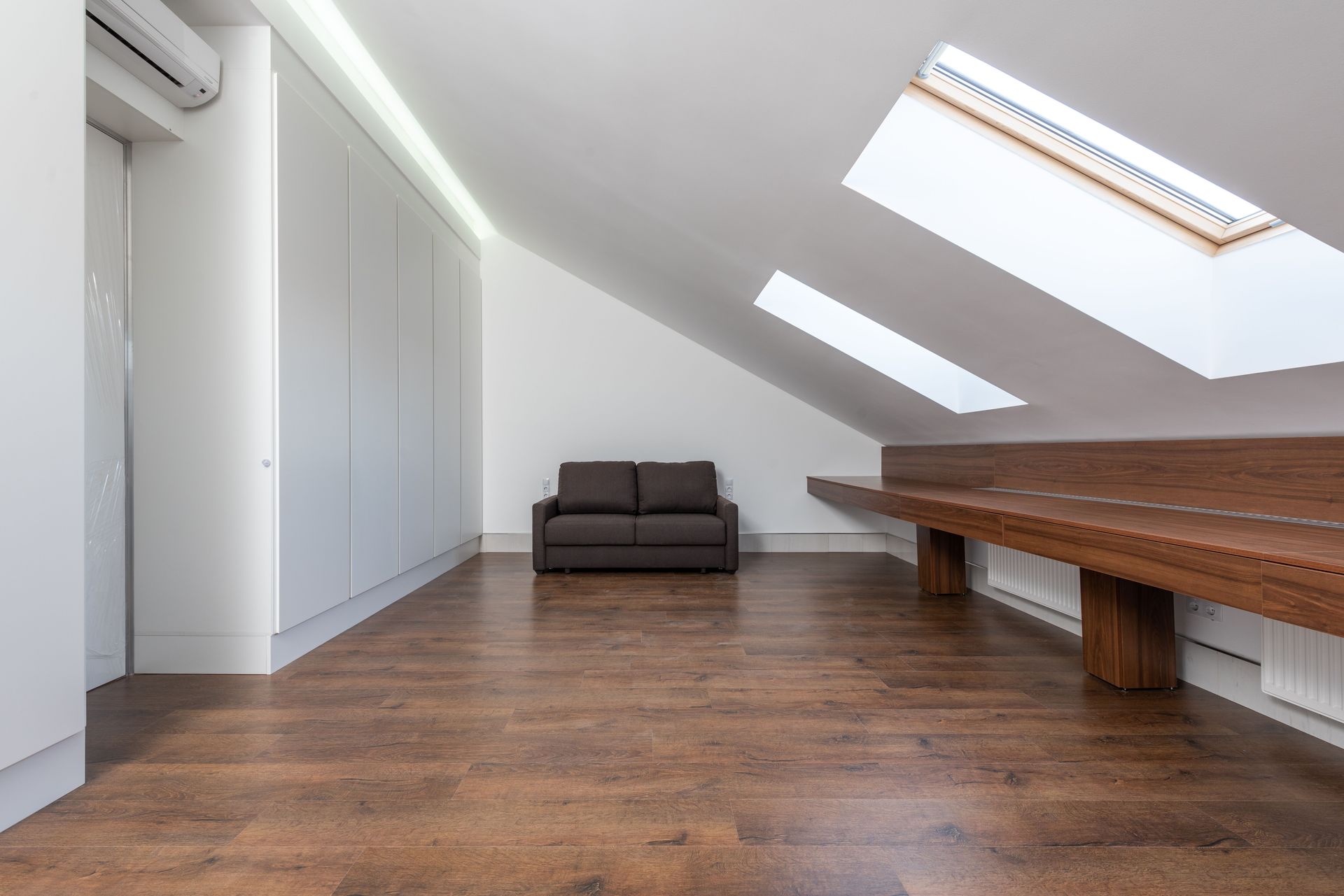 Spacious attic room featuring large skylights, a brown sofa, and wooden flooring.